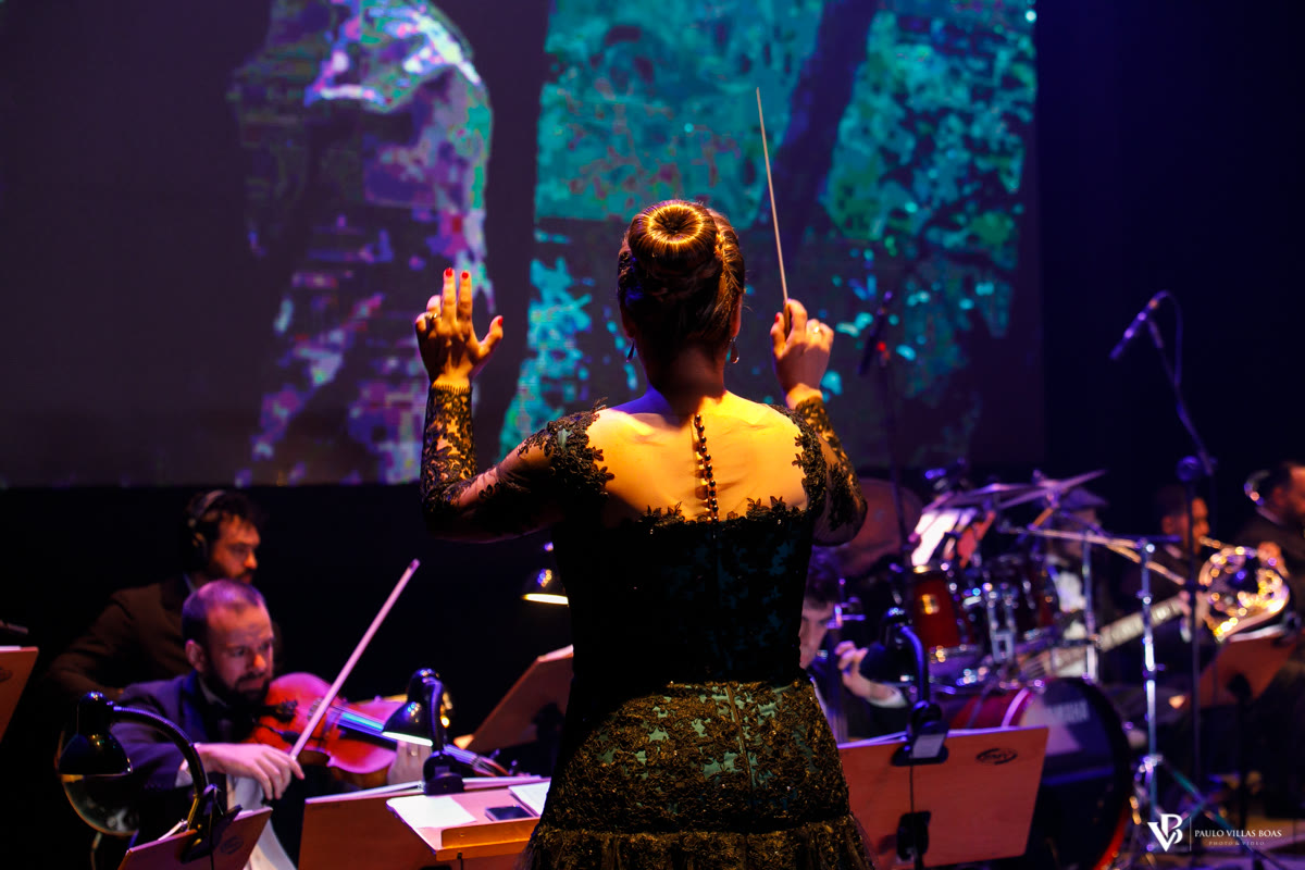 Marianna Ruggiero de costas conduzindo músicos em uma apresentação